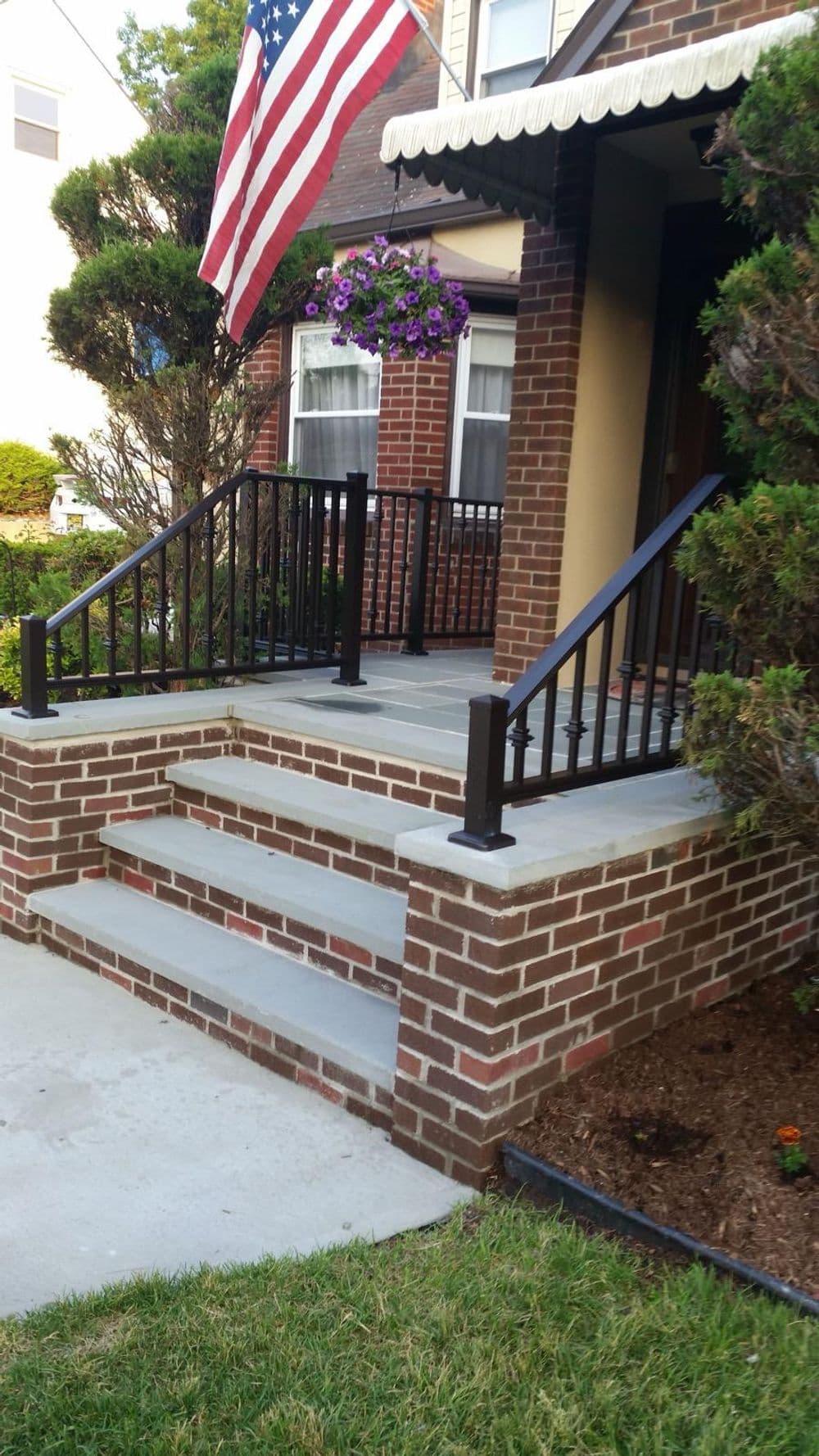 Front porch with brick steps, black railing, and American flag, surrounded by greenery.