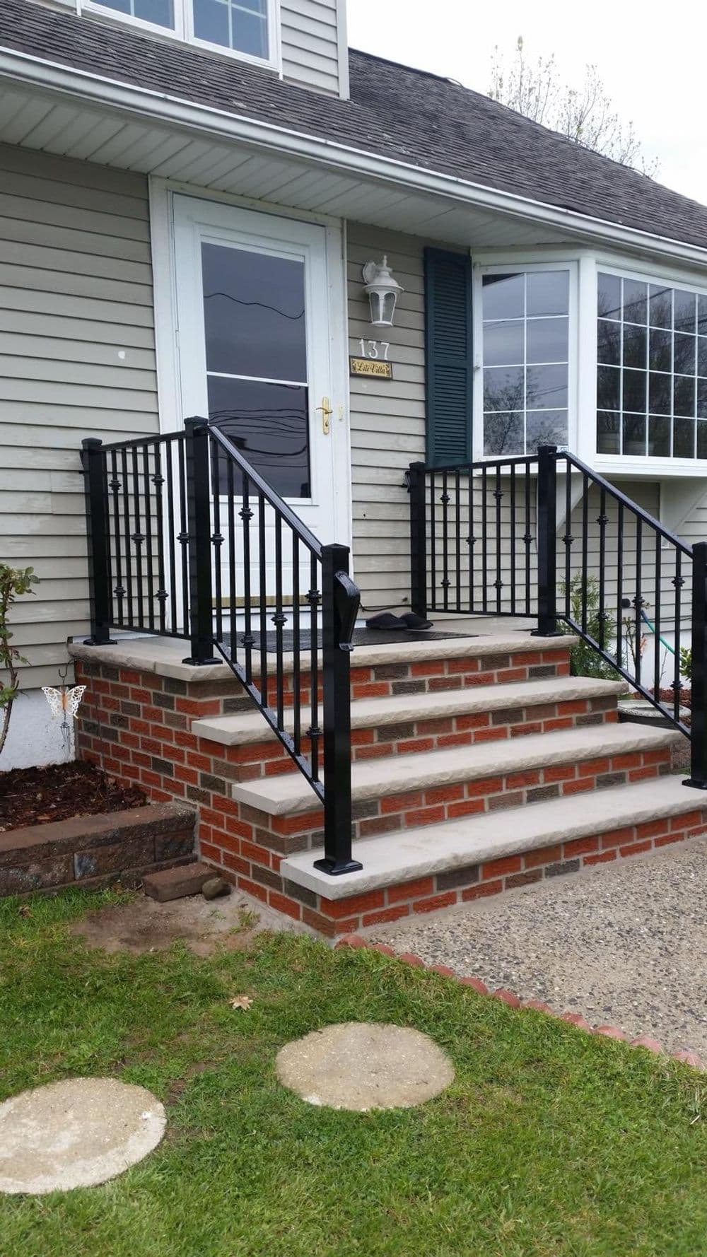 Front entrance with black railing and brick steps leading to a home. Green lawn and window view.