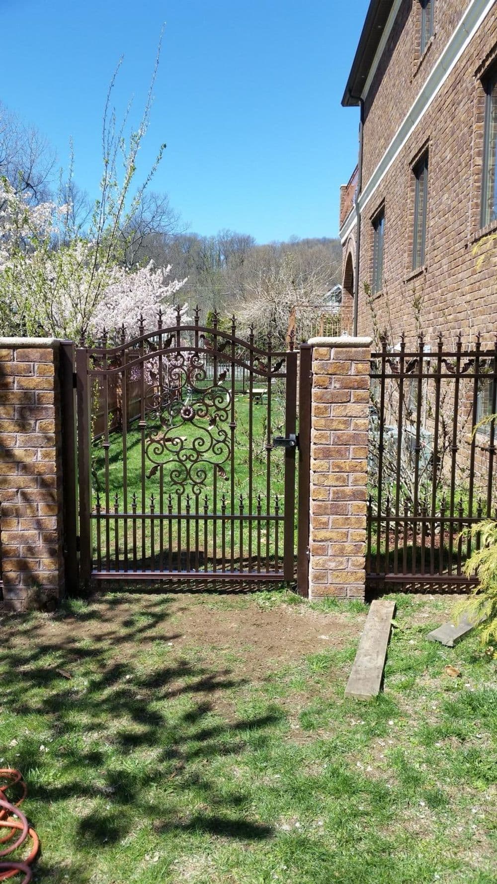 Iron gate with decorative design leading to a lush green garden and blooming trees.