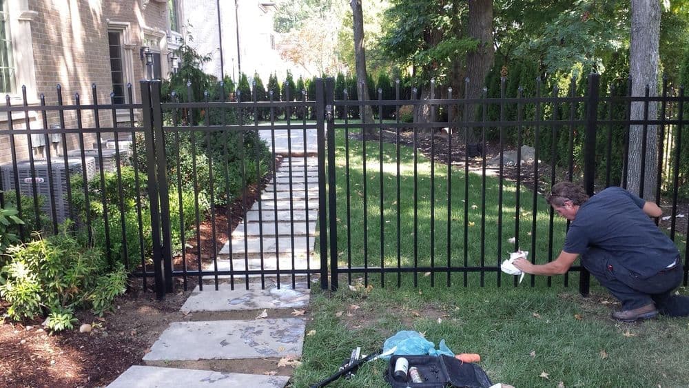 Man performs maintenance on a black metal fence in a landscaped yard with a stone pathway.