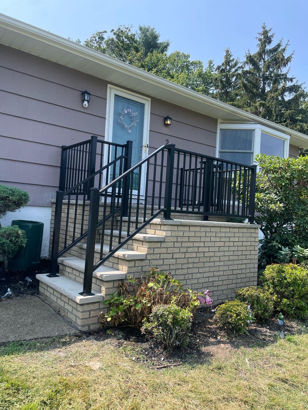 Front entrance of a house with stairs and black railings, surrounded by greenery.