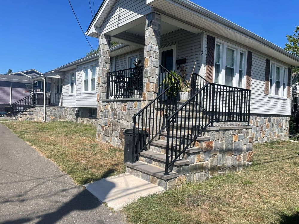 Stone front steps lead to a charming gray house with a porch and lush landscaping.
