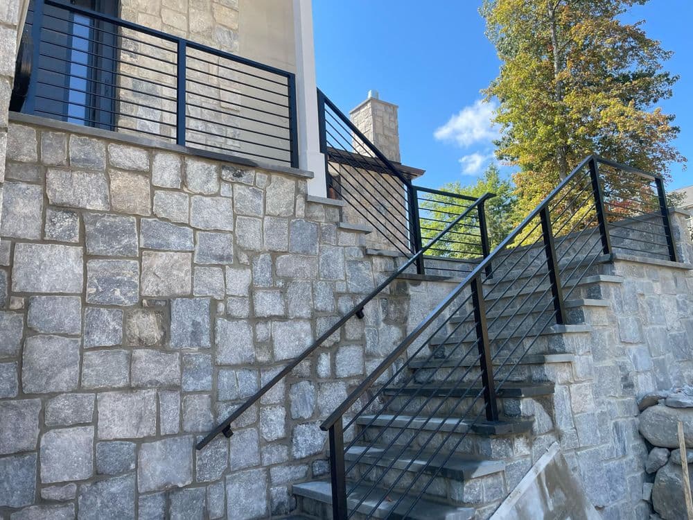 Modern stone staircase with black metal railing and stone walls under a bright blue sky.