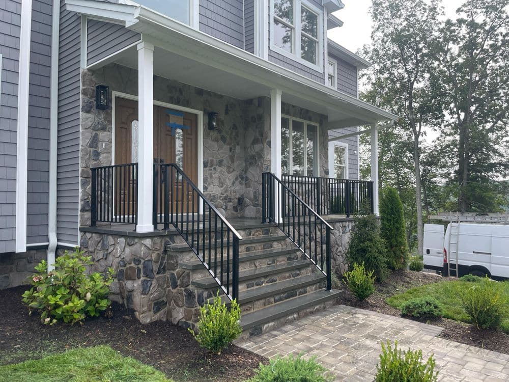 Modern home entrance with stone steps, black railing, and landscaped surroundings.