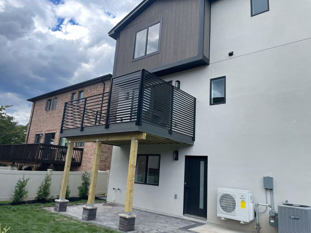 Modern home exterior featuring a balcony, patio, and HVAC unit with cloudy sky backdrop.
