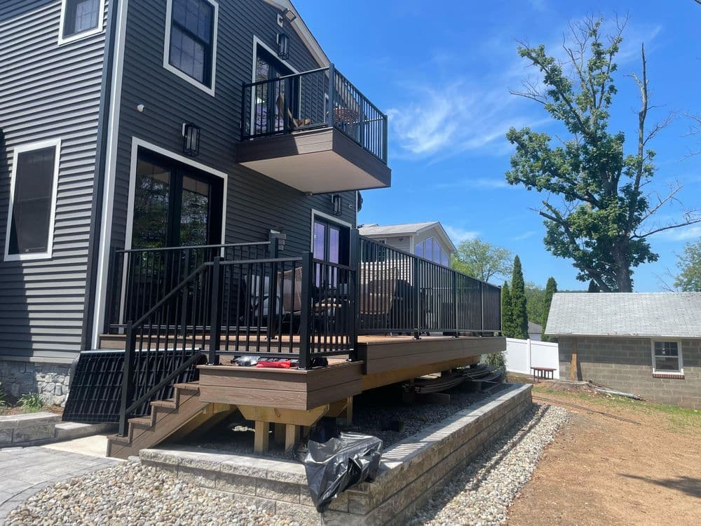 Modern home exterior featuring a wood deck, black railing, and greenery under a clear blue sky.