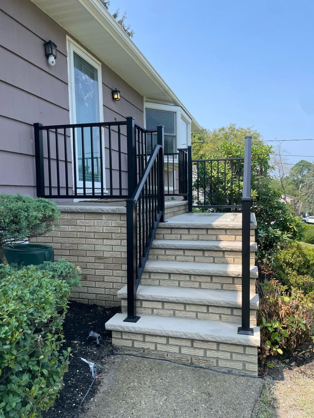 Black metal railing on a brick staircase leading to a home entrance surrounded by greenery.
