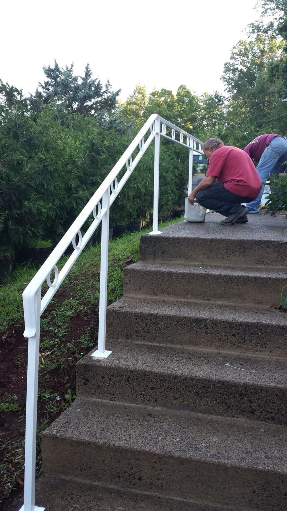 Person installing a white railing on outdoor concrete steps surrounded by greenery.