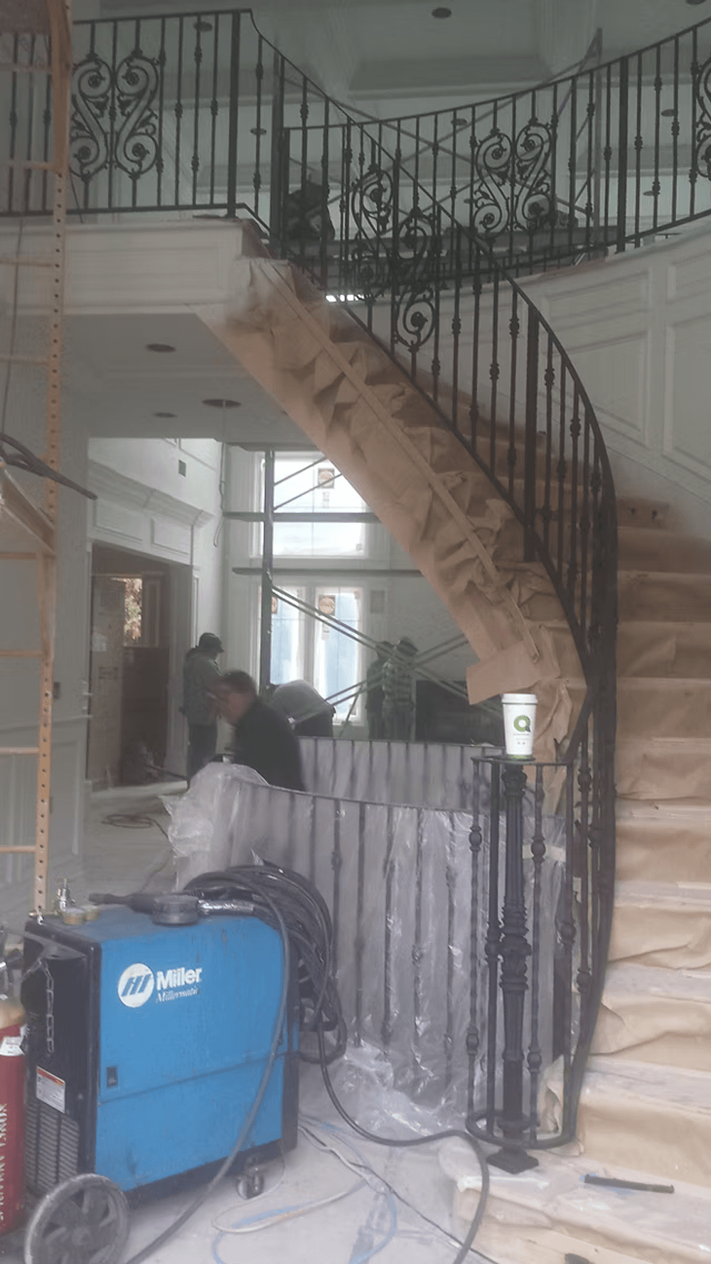 Curved staircase under renovation, featuring black iron railing and construction equipment nearby.