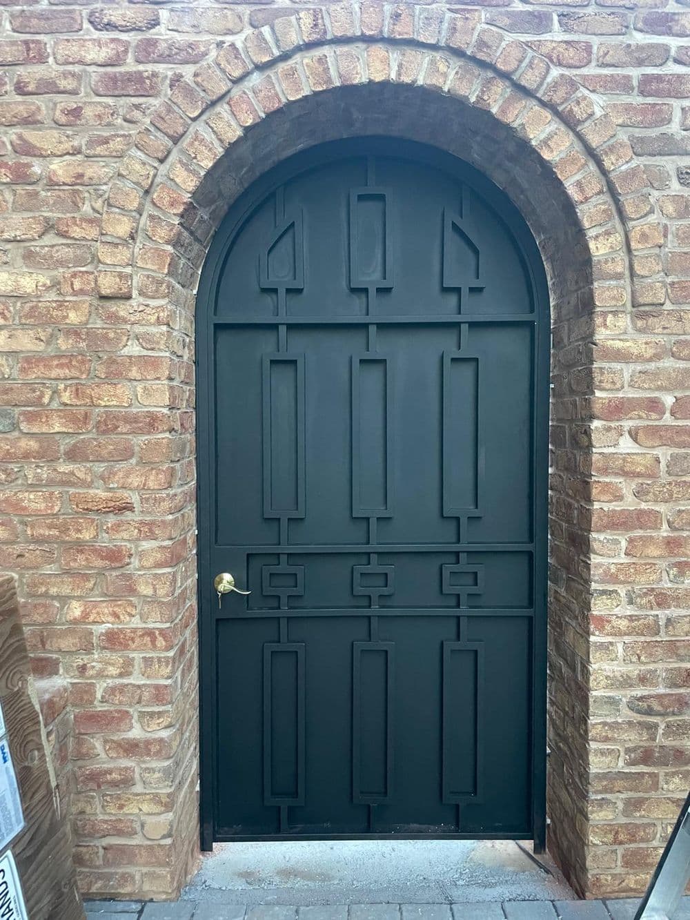 Arched black wrought iron door with geometric design and brick wall background.