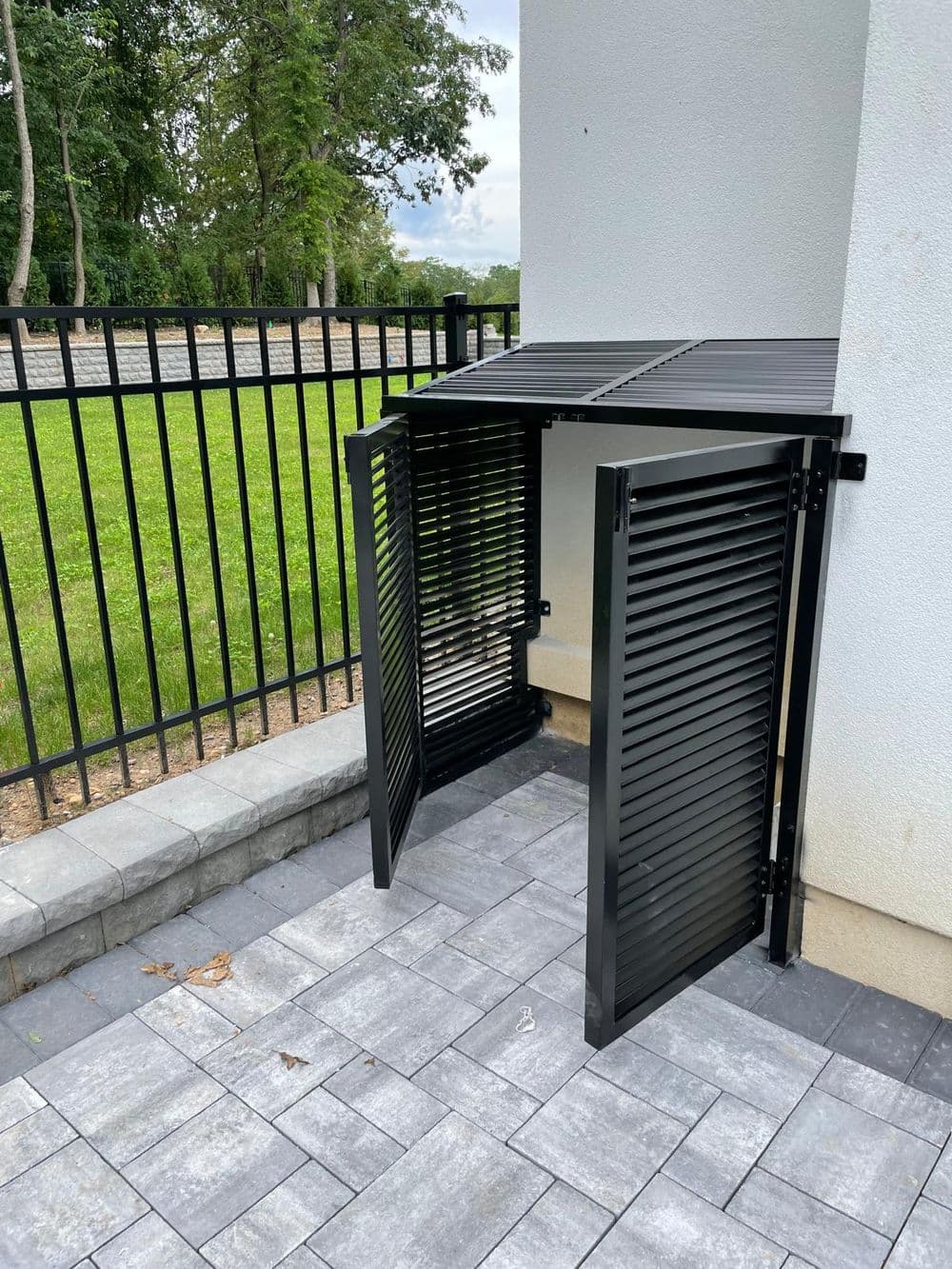 Black metal storage shed with slatted design near a fence on a stone patio.