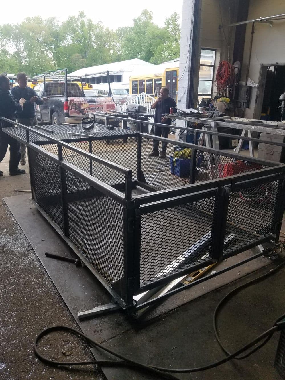 Workers assembling a metal mesh cage in a workshop, with vehicles in the background.