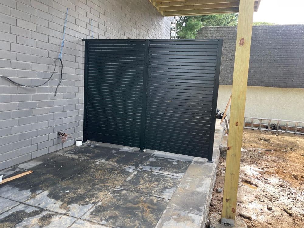 Modern black slatted fence installed beside a gray brick wall on a concrete patio.