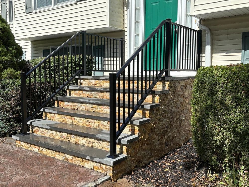 Black metal railing on stone steps leading to a green door entrance. Landscaping in view.