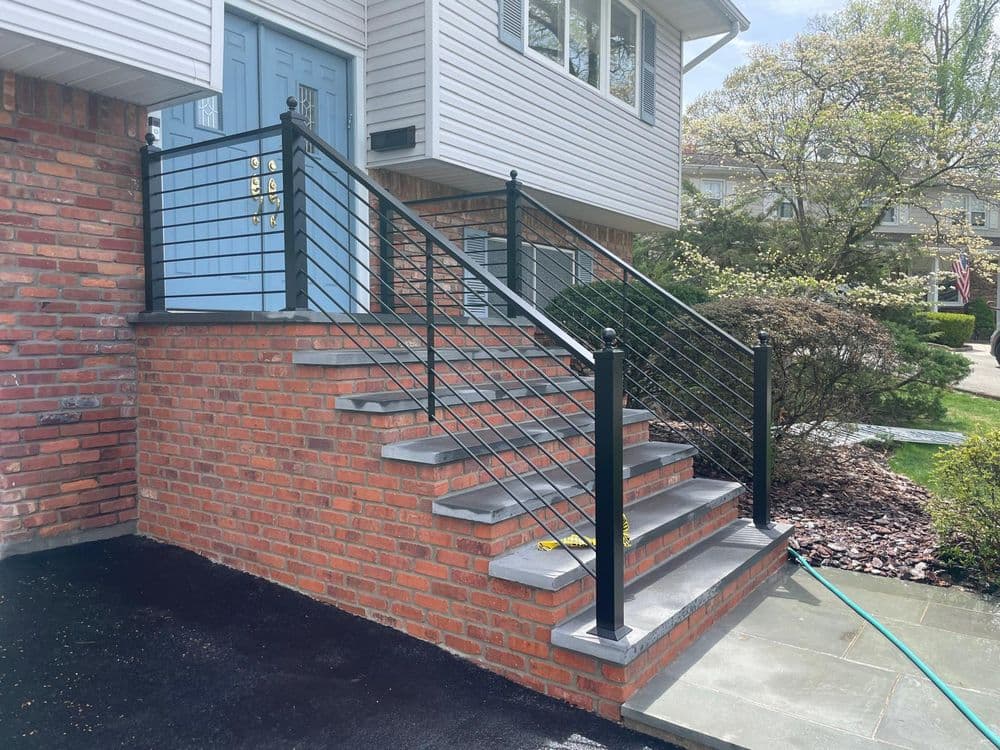 Modern brick staircase with sleek metal railing leads to a home entrance. Bright, well-maintained landscaping.