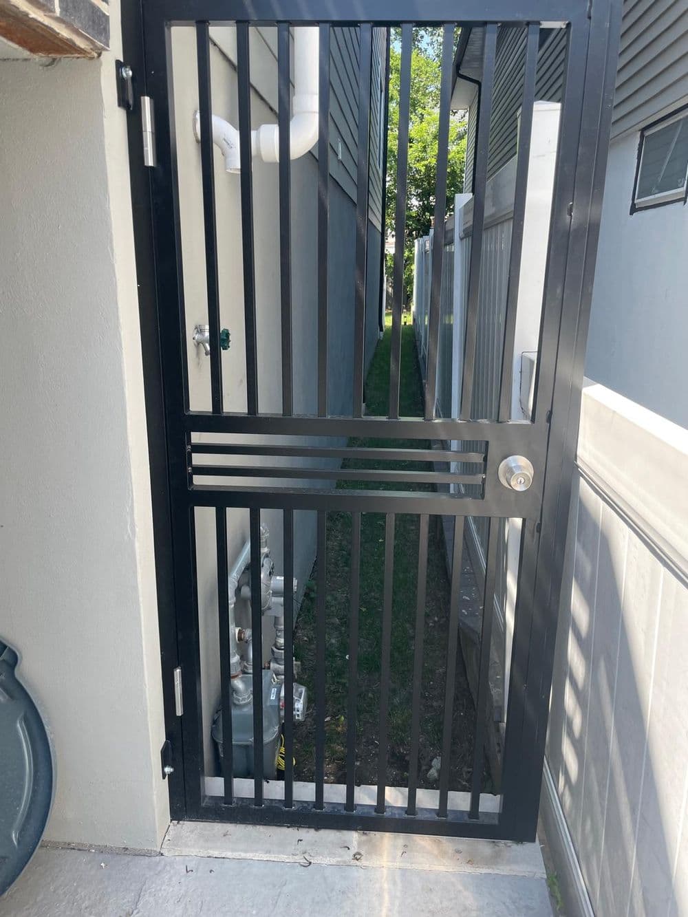 Black metal gate leading to narrow side yard between buildings, with visible utilities.