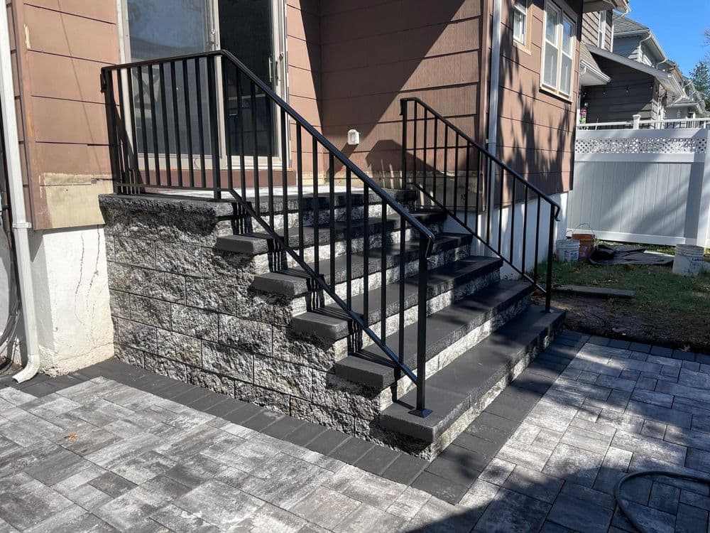 Black metal railing on stone steps leading to a house, surrounded by a paved patio.