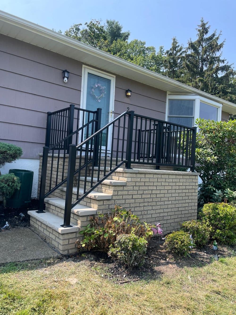 Front entrance of a house with brick steps and a black railing, surrounded by greenery.