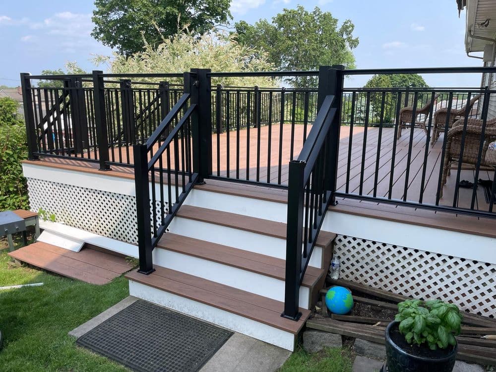 Modern outdoor deck with black railing, steps, and lush greenery in the background.
