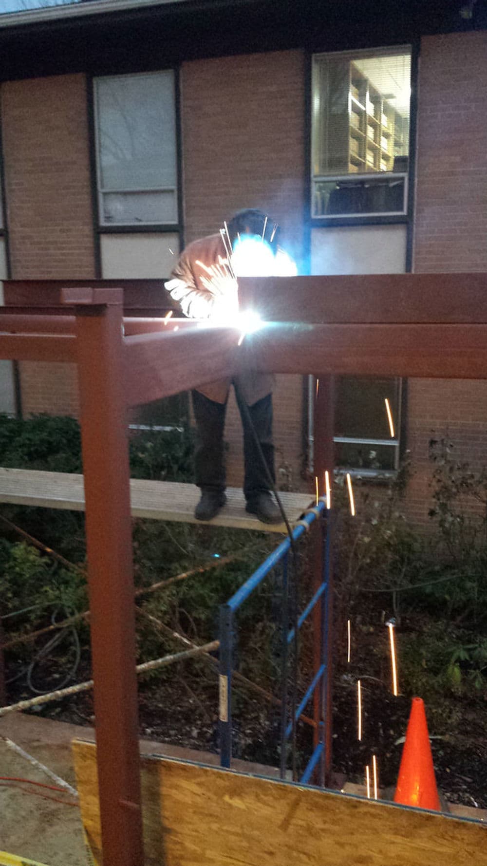 Welder working on steel structure, sparks flying, with scaffolding and a safety cone nearby.