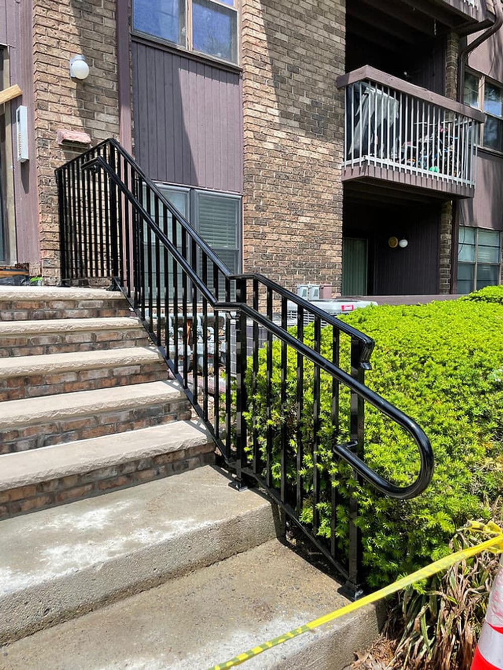 Black handrails on exterior stairs leading to an apartment building, surrounded by green shrubs.