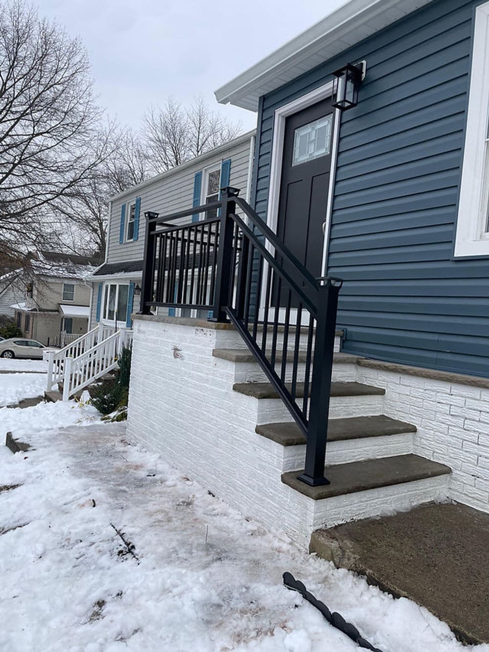 Black railing on steps leading up to a blue house in winter with snow on the ground.