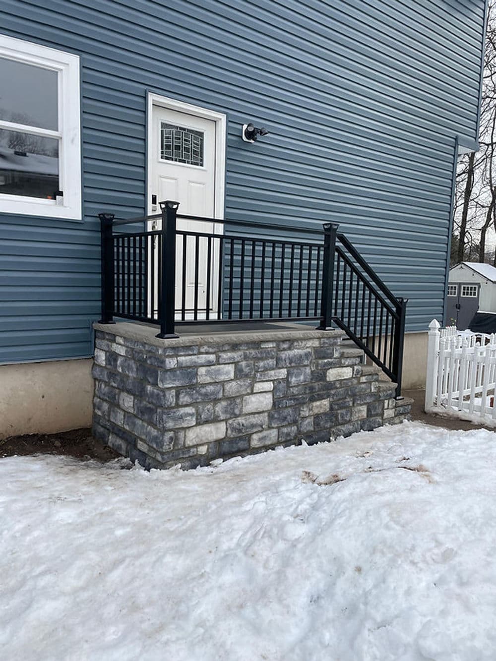 Exterior stone steps with black railing leading to a white door on a blue house. Snow on the ground.