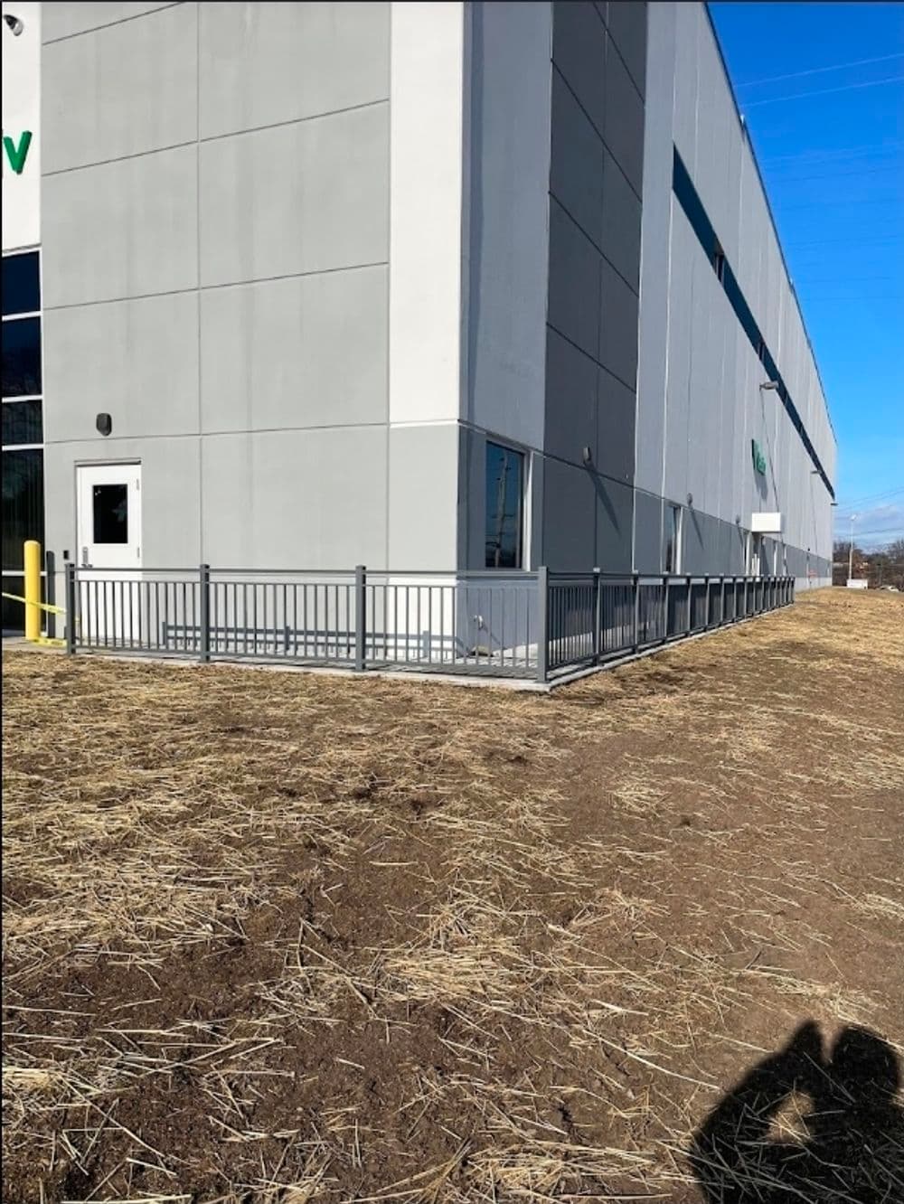 Gray building corner with fenced area and barren ground, bright blue sky above.