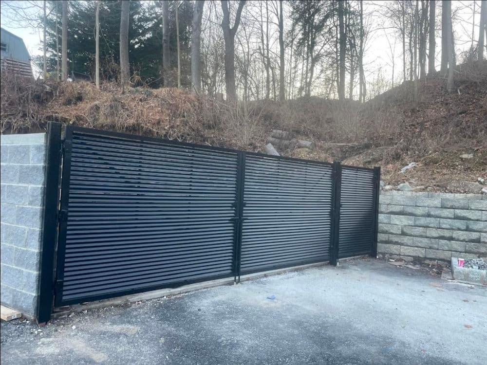 Black sliding security gate in a gravel area surrounded by trees and retaining wall.