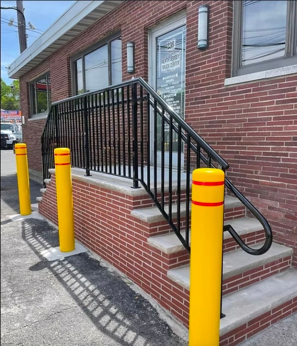Exterior view of a brick building with yellow safety poles and a black railing on stairs.