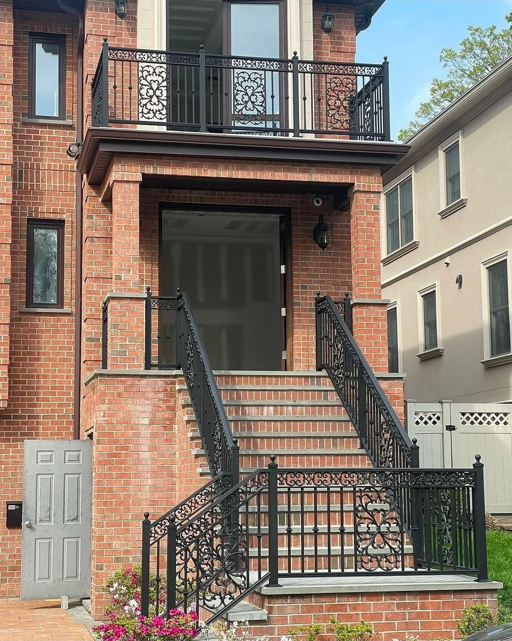 Elegant brick home entrance with black iron railing and stairs, surrounded by greenery.