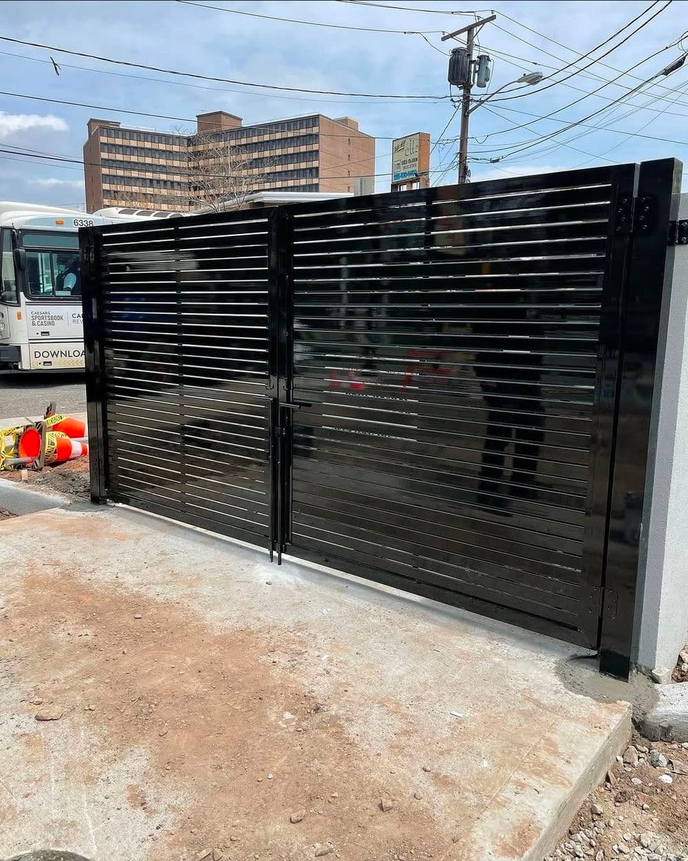 Black metal gate with horizontal slats, installed on a concrete base near urban landscape.