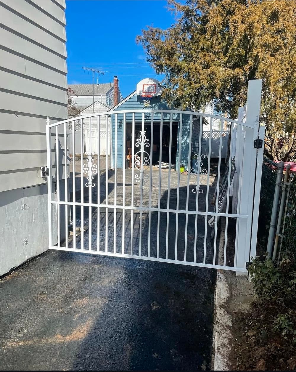White decorative driveway gate with elegant patterns, leading to a paved area and garage.