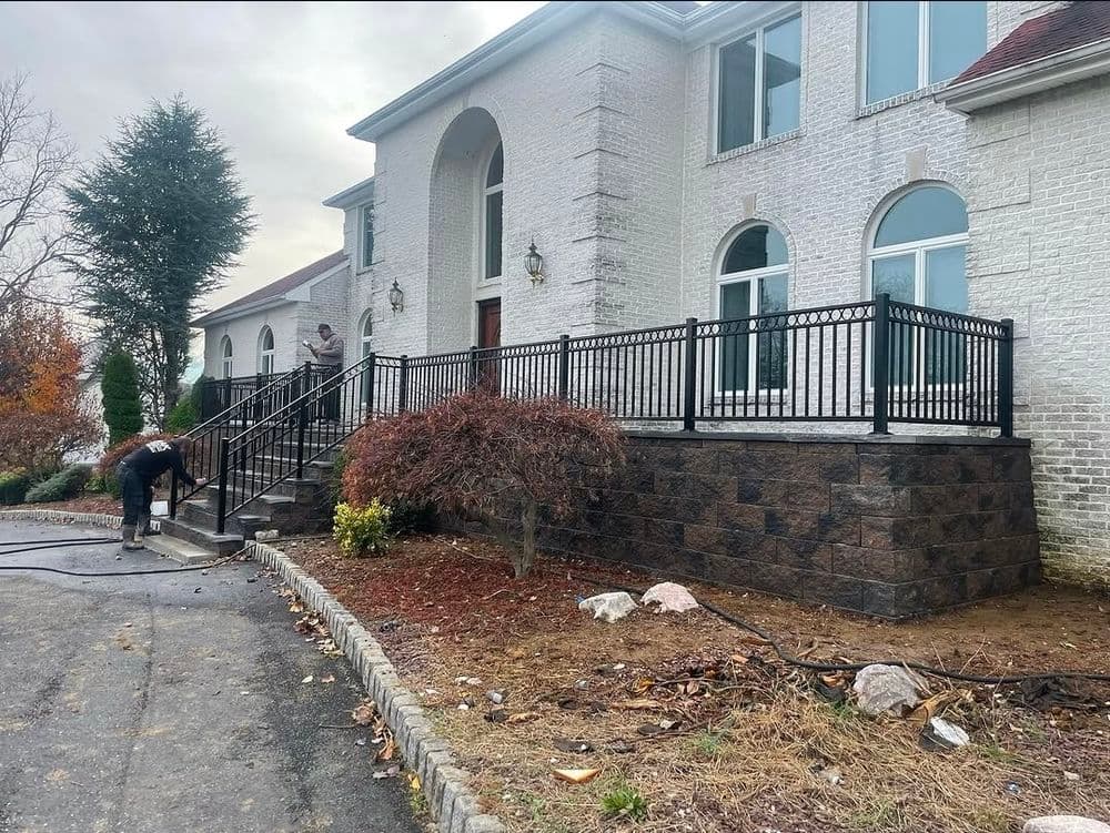 Exterior of a white brick house with a black railing, landscaping, and workers on the steps.