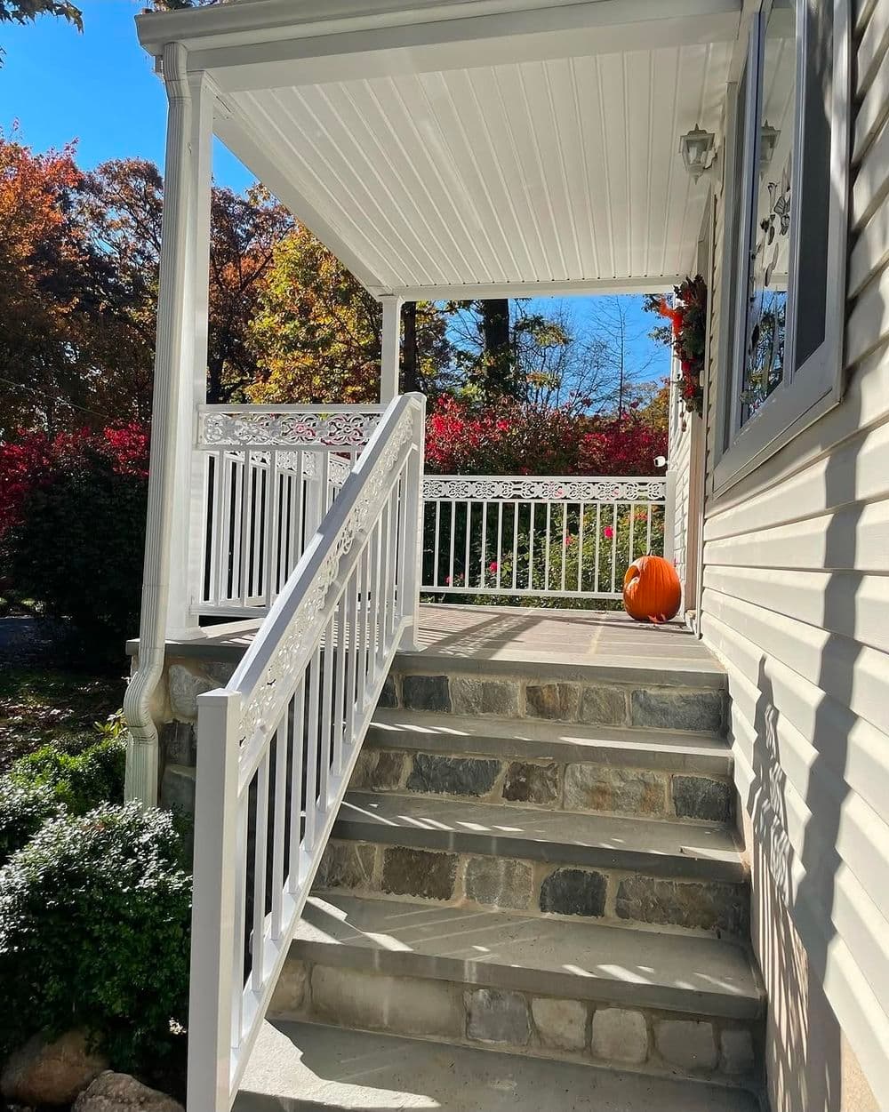 White porch with stairs, decorative railings, and a pumpkin in autumn garden setting.
