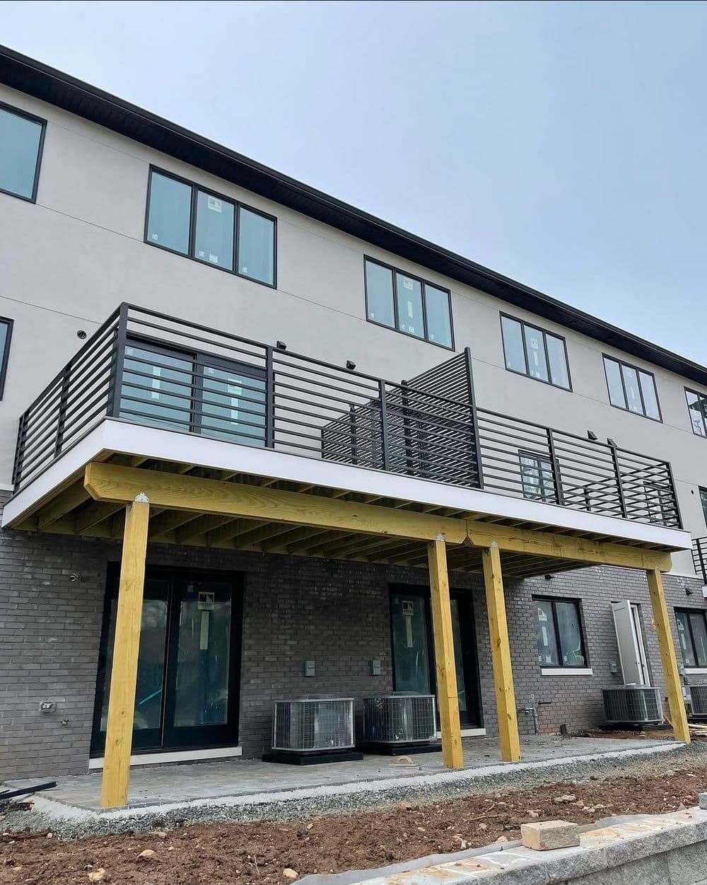 Modern two-story building with a wooden deck, metal railings, and air conditioning units below.