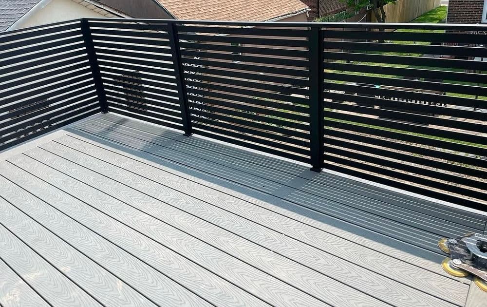 Modern outdoor deck with sleek black railing and textured gray wood planks, sunlight casting shadows.