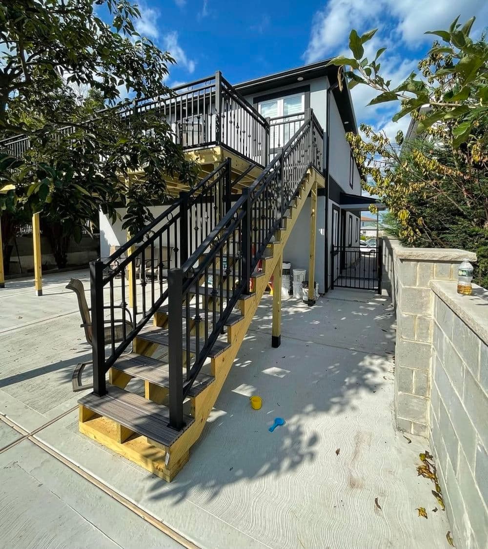 Wooden staircase with black railings leading to a modern house on a sunny day.
