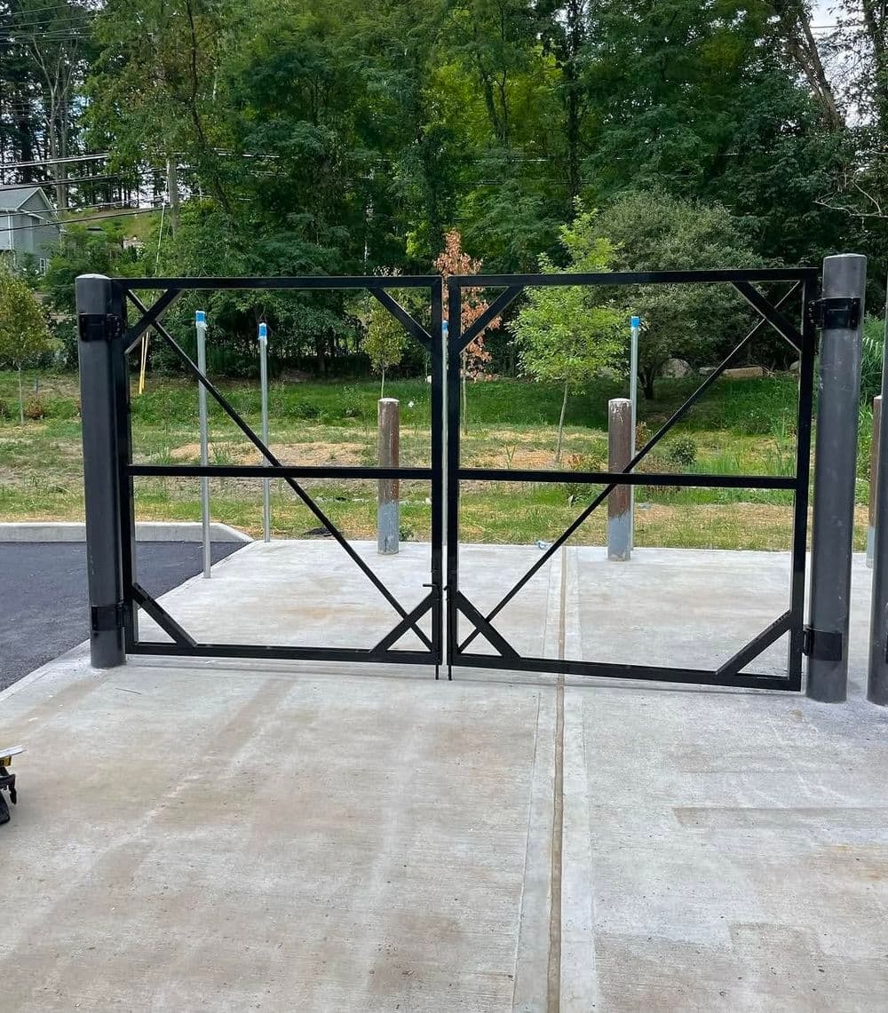 Black metal gate with cross-bracing, surrounded by greenery and concrete pavement.