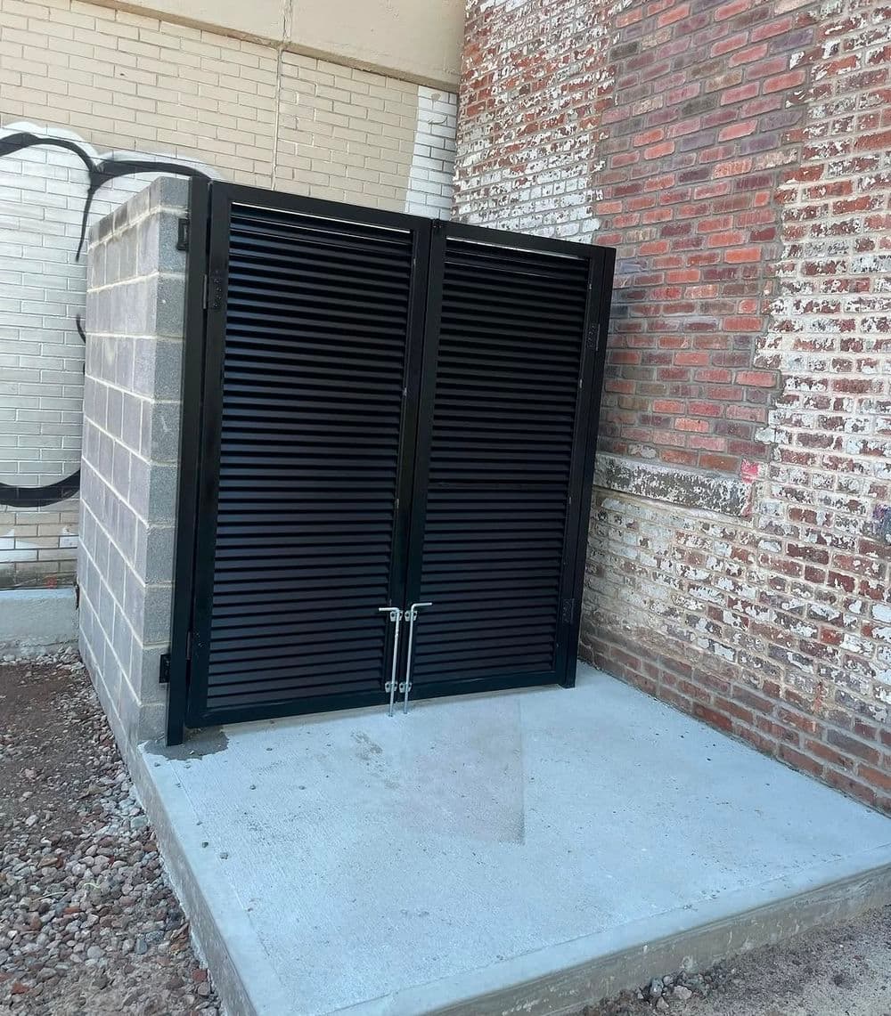 Black metal storage shed with louvered doors against a brick wall. Durable and secure.