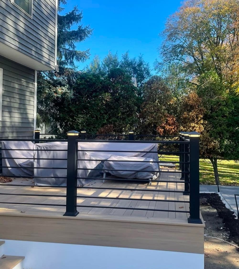 Deck with modern railing and covered furniture, surrounded by greenery under blue sky.