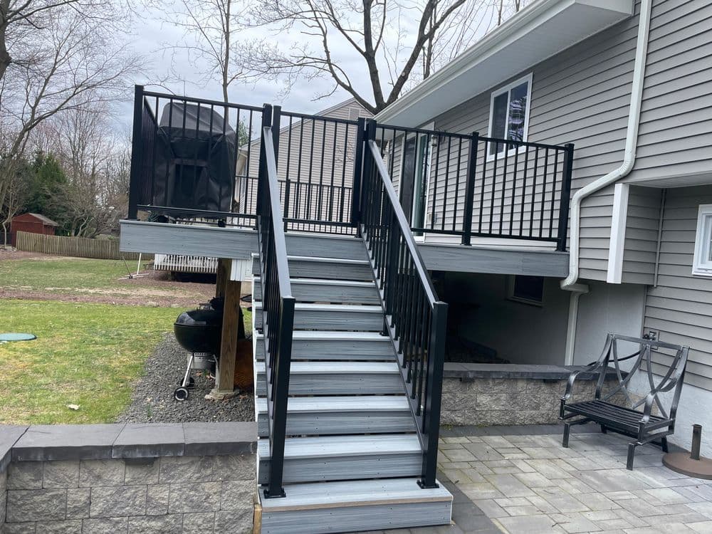 Modern wooden deck with black railings and stairs leading to a house. Grassy yard in background.