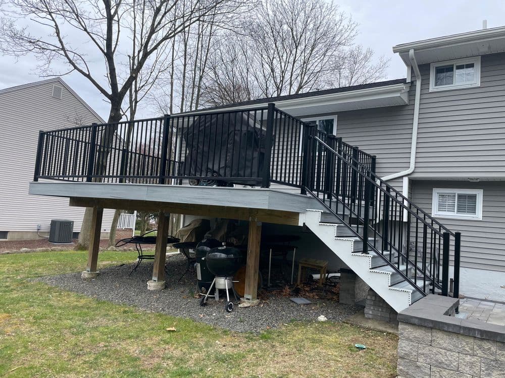 Elevated deck with black railing and stairs, surrounded by grass and trees, residential area.