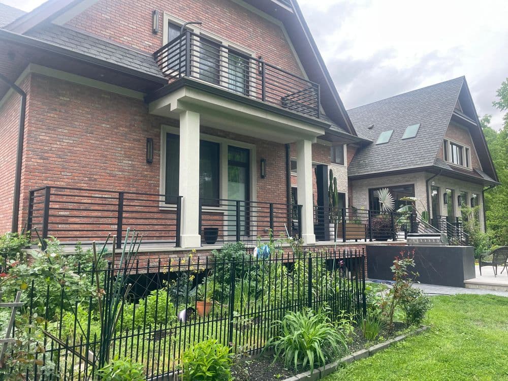 Modern brick house with stylish balconies, surrounded by a lush garden and fencing.