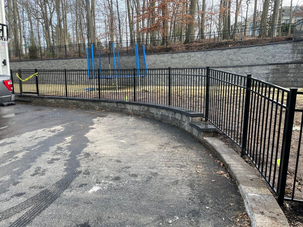 Black metal fence surrounding a curved stone wall with a trampoline in the background.