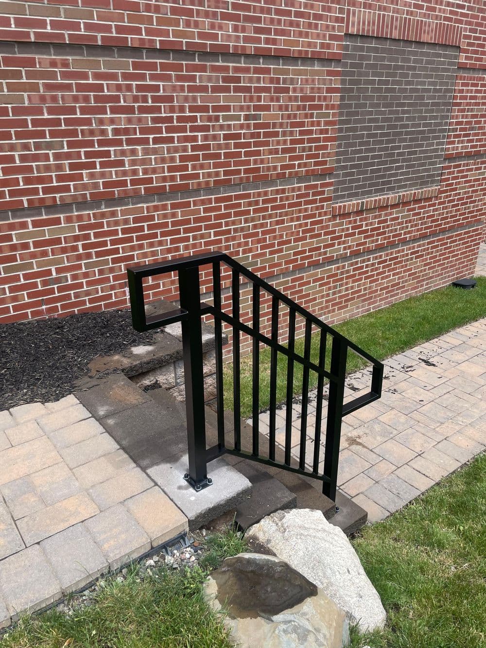 Black metal handrail on brick building stairs with stone pathway and grass.