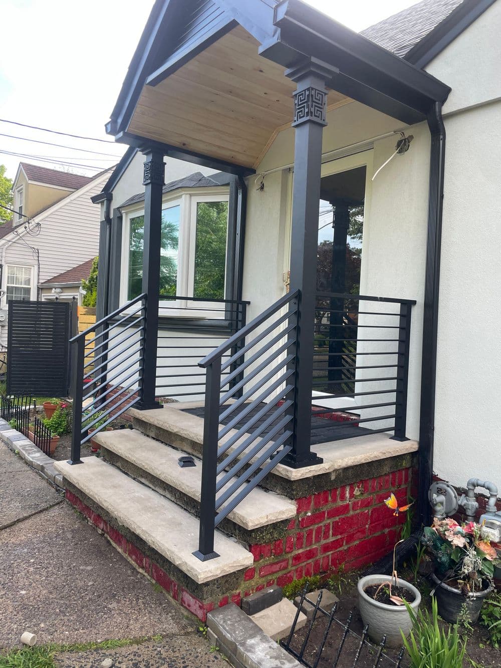 Modern front porch with black railings and stone steps, featuring a wooden overhang and plants.