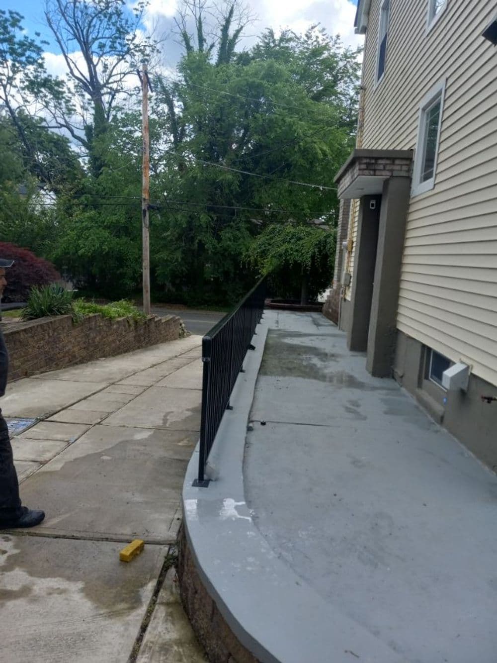Concrete walkway with black railing beside a house, surrounded by greenery.