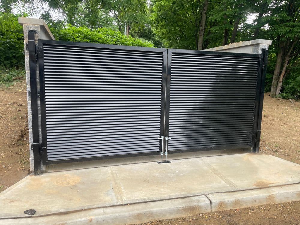 Modern black horizontal slatted gate on a concrete base surrounded by green foliage.