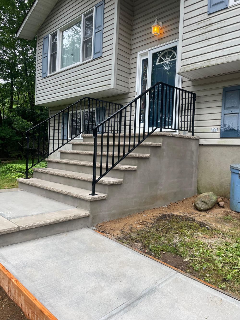 Residential entrance with steps, black railing, and new concrete walkway. Green landscaping.
